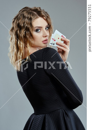 Blonde woman in black dress showing two playing cards, posing sideways against gray background. Gambling entertainment, poker, casino. Close-up. Blonde woman in black dress showing two playing cards, posing sideways against gray background. Gambling entertainment, poker, casino. Close-up. 79275551