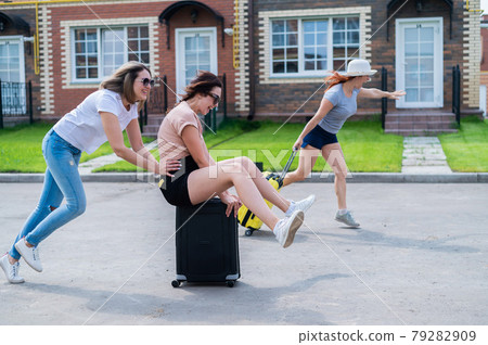 Three joyful Caucasian friends are having fun and chasing each other while riding a suitcase. Excited women go on a road trip. The brunette blonde and the redhead are going on vacation.  79282909