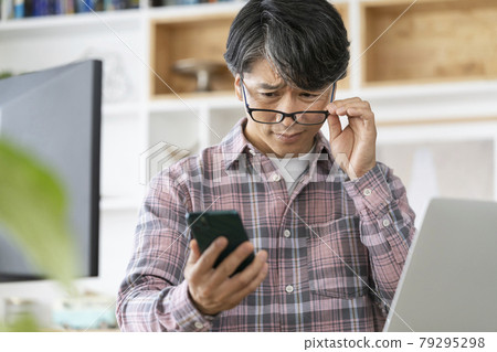 A man in his 50s looking at the screen of his smartphone by shifting his reading glasses A man in his 50s looking at the screen of his smartphone by shifting his reading glasses 79295298