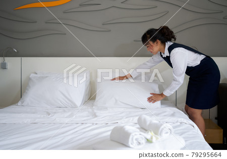 Young hotel maid making the bed. Hotel staff in blue uniform preparing room for guest. 79295664