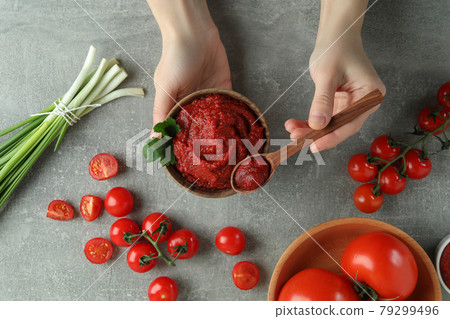 Female hands hold bowl and spoon with tomato paste on gray background with ingredients 79299496