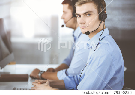 Two young men in headset, sitting at the desk in a sunny modern office, listening to the clients. Call center operators at work 79300236