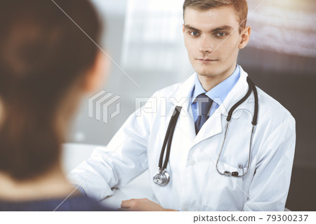 Doctor man, with a stethoscope, is listening to his patient, while sitting at the desk in the sunny cabinet at hospital. Perfect medical service in clinic. Happy future in medicine and healthcare Doctor man, with a stethoscope, is listening to his patient, while sitting at the desk in the sunny cabinet at hospital. Perfect medical service in clinic. Happy future in medicine and healthcare 79300237