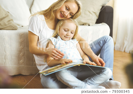 Happy family. Blonde young mother reading a book to her cute daughter while sitting at wooden floor. Motherhood concept 79300345