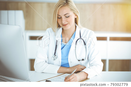 Woman-doctor at work while sitting at the desk in sunny clinic. Blonde cheerful physician filling up medication history record form 79302779