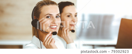 Blond female customer service representative is consulting clients online using headset in sunny office. Call center concept Blond female customer service representative is consulting clients online using headset in sunny office. Call center concept 79302923