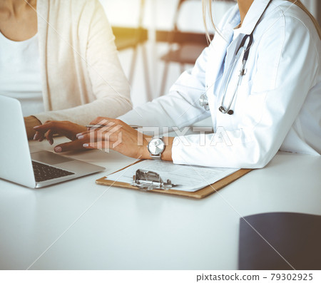 Unknown woman-doctor communicating with female patient in sunny clinic. Physician checks medical history record and exam results, close-up Unknown woman-doctor communicating with female patient in sunny clinic. Physician checks medical history record and exam results, close-up 79302925