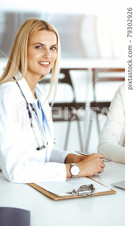 Woman-doctor at work in sunny hospital is happy to consult female patient. Blonde physician checks medical history record and exam results while using clipboard Woman-doctor at work in sunny hospital is happy to consult female patient. Blonde physician checks medical history record and exam results while using clipboard 79302926