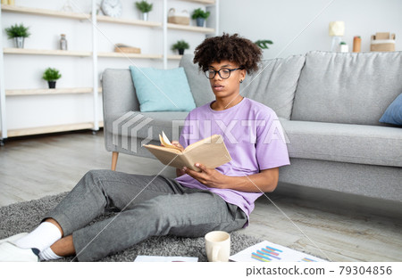 Focused black teenager reading book, sitting on floor, studying from home 79304856