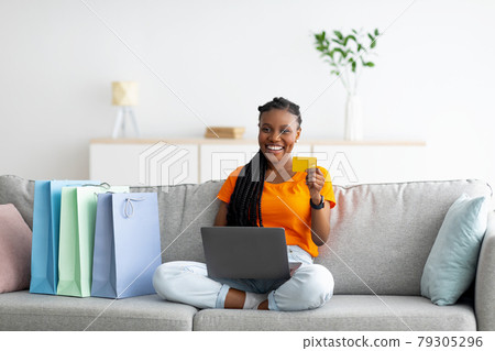 Cheerful black woman sitting on couch with laptop computer, holding credit card, making payment in online shop from home Cheerful black woman sitting on couch with laptop computer, holding credit card, making payment in online shop from home 79305296