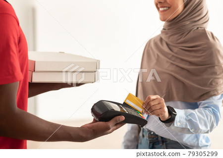 Deliveryman holding POS machine, muslim woman paying with card 79305299