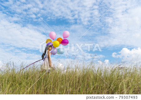 Cheerful cute girl holding balloons running on green meadow white cloud and blue sky with happiness. Hands holding vibrant air balloons play on birthday party happy times summer on sunlight outdoor 79307493