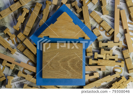 A wooden house on a blue lining on top of a wooden puzzle and money. 79308046