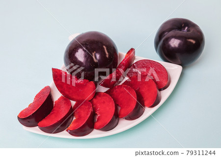 On a white saucer there is a large plum cut into wedges, next to it there are two more plums. On a white saucer there is a large plum cut into wedges, next to it there are two more plums. 79311244