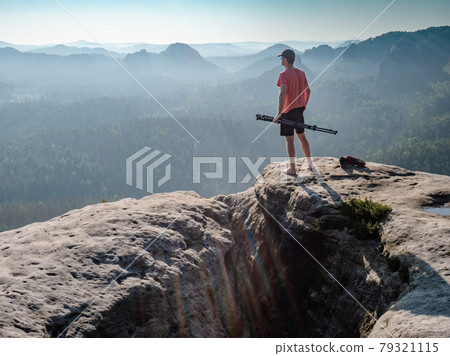 Barefoot man in red t-shirt stay with tripod in hand on rocky viewpoint and looking Barefoot man in red t-shirt stay with tripod in hand on rocky viewpoint and looking 79321115
