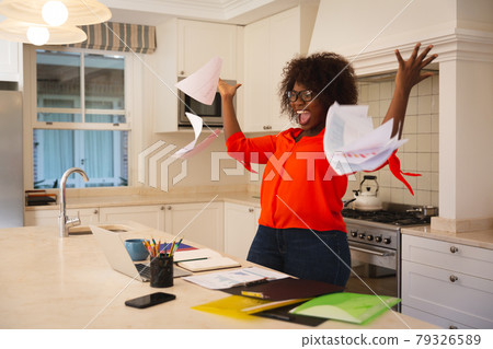 Happy african american woman working in kitchen with laptop, celebrating success, throwing papers 79326589