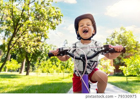 Portrait little cute adorable caucasian toddler boy in safety helmet enjoy having fun riding exercise bike in city park road yard garden forest. Child first bike. Kid outdoors sport summer activities Portrait little cute adorable caucasian toddler boy in safety helmet enjoy having fun riding exercise bike in city park road yard garden forest. Child first bike. Kid outdoors sport summer activities 79330975