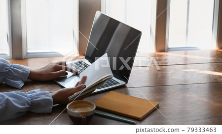 Side view of college student is reading book to find information and using laptop on wooden desk. Side view of college student is reading book to find information and using laptop on wooden desk. 79333463