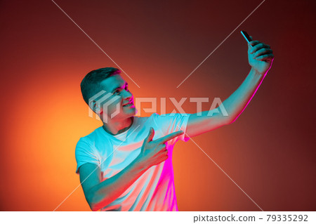 Portrait of young Caucasian man using phone isolated over gradient red orange studio background in neon light with copyspace for ad. Concept of human emotions 79335292