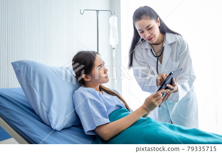 Asian female doctor therapeutic advising with tablet to young female patient on Bed in hospital background. 79335715