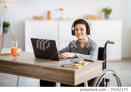 Home schooling. Cheerful disabled teenager in wheelchair wearing headphones, having online lesson on laptop indoors Home schooling. Cheerful disabled teenager in wheelchair wearing headphones, having online lesson on laptop indoors 79342375