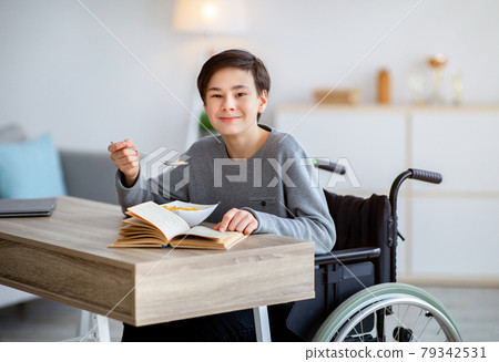 Cool disabled teenager in wheelchair having cereal for breakfast and reading book at home. Hobbies and pastimes Cool disabled teenager in wheelchair having cereal for breakfast and reading book at home. Hobbies and pastimes 79342531