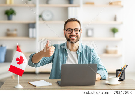 Happy young guy sitting at table with flag of Canada, using laptop computer, showing thumb up gesture at home Happy young guy sitting at table with flag of Canada, using laptop computer, showing thumb up gesture at home 79342898