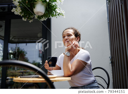 Charming woman sits in a cafe on a summer terrace and looks thoughtfully into the distance with a cup of coffee in her hand Charming woman sits in a cafe on a summer terrace and looks thoughtfully into the distance with a cup of coffee in her hand 79344151