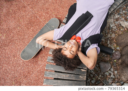 Guy laying at the bench with closed eyes and relaxing after riding at the skateboard Guy laying at the bench with closed eyes and relaxing after riding at the skateboard 79365314