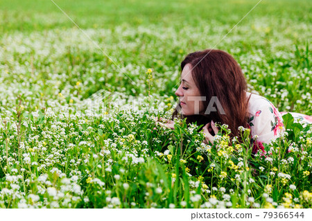 Beautiful brunette woman in dress with flowers sits in spring field Beautiful brunette woman in dress with flowers sits in spring field 79366544
