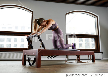 A woman yoga instructor trains on a reformed Cadillac in a large hall against a window, modern equipment for training in gyms, correction of the musculoskeletal system. 79367348