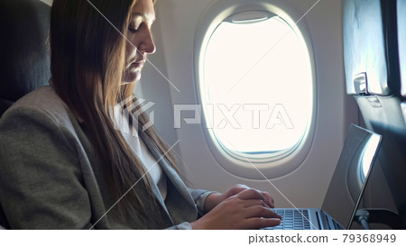 Young woman in a suit typing text on a laptop at the window in an airplane 79368949
