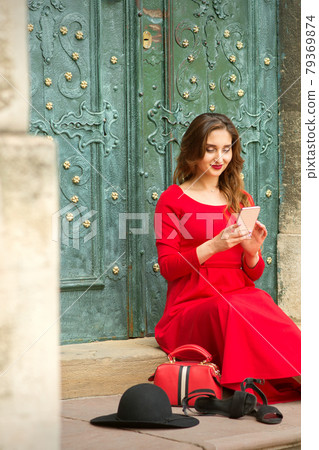 Beautiful young caucasian woman sitting on the stairs by the door with travel suitcase and smartphone wearing long red dress. Beautiful young caucasian woman sitting on the stairs by the door with travel suitcase and smartphone wearing long red dress. 79369874
