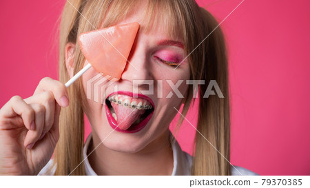 Portrait of a young woman with braces and bright makeup eating a lollipop on a pink background. 79370385