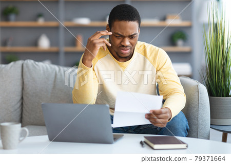 Confused black man sitting in front of laptop, holding papers 79371664