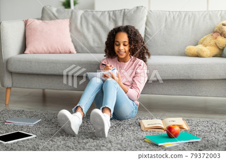 Positive african american teen girl doing homework, making notes or writing essay, sitting in living room interior Positive african american teen girl doing homework, making notes or writing essay, sitting in living room interior 79372003