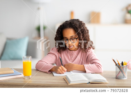 Cute african american schoolgirl in glasses writing in notepad, doing homework or writing essay, studying at home 79372828
