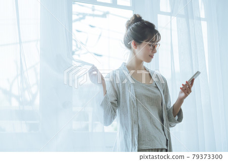 一位身著年輕家居服的女性在窗前看著智能手機的生活方式形象 一位身著年輕家居服的女性在窗前看著智能手機的生活方式形象 79373300