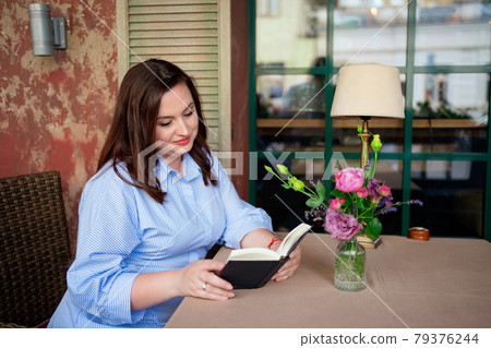 a woman sits at a table in a cafe or on the veranda and reading a book aloud. a woman sits at a table in a cafe or on the veranda and reading a book aloud. 79376244