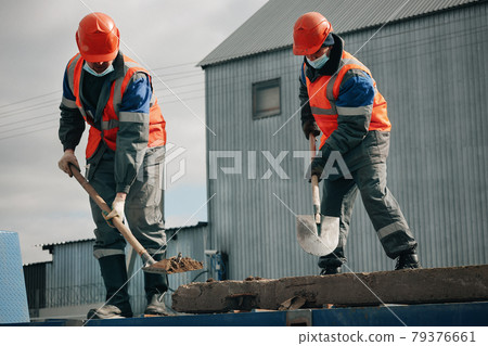 Two workers in hard hats, work clothes and a medical mask work with shovels at a construction site Two workers in hard hats, work clothes and a medical mask work with shovels at a construction site 79376661