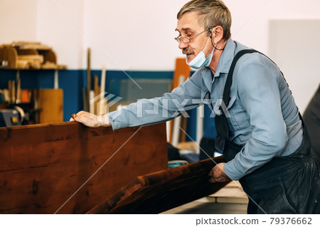A gray-haired carpenter with glasses works with wood on a workbench in a carpentry shop. A gray-haired carpenter with glasses works with wood on a workbench in a carpentry shop. 79376662
