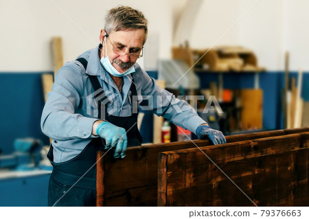An elderly man paints wooden boards with dark varnish in a carpentry workshop. A pensioner in a An elderly man paints wooden boards with dark varnish in a carpentry workshop. A pensioner in a 79376663
