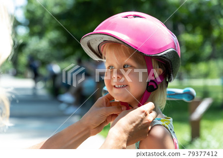 Close-up mom parent hand put on and fasten safety helmet on cute blond caucasian toddler girl for riding bike or scooter city street park outdoors on summer day. Child sport activity protection care Close-up mom parent hand put on and fasten safety helmet on cute blond caucasian toddler girl for riding bike or scooter city street park outdoors on summer day. Child sport activity protection care 79377412