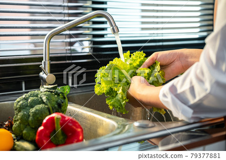 Asian housewife wearing an apron and standing to washing lettuce and vegetables with water in sink 79377881