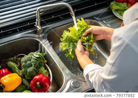 Asian housewife wearing an apron and standing to washing lettuce and vegetables with water in sink 79377882