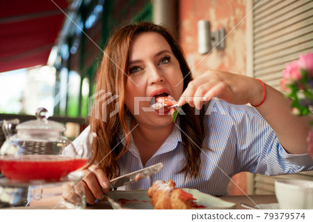 gluttony. A funny woman eats a croissant with strawberries with a knife and fork gluttony. A funny woman eats a croissant with strawberries with a knife and fork 79379574