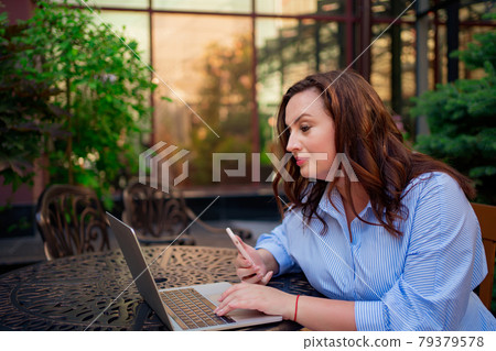 The woman sits on an outdoor terrace and works on a laptop and a smartphone. The woman sits on an outdoor terrace and works on a laptop and a smartphone. 79379578