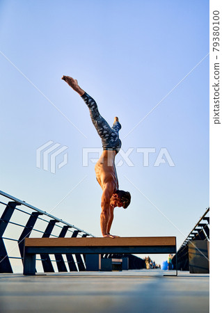 Fit man doing a hand stand while practicing yoga alone near the ocean against sky at dusk or dawn 79380100