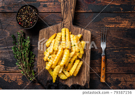 Baked Crinkle French fries potatoes sticks or chips on a wooden board. Dark wooden background. Top view Baked Crinkle French fries potatoes sticks or chips on a wooden board. Dark wooden background. Top view 79387156