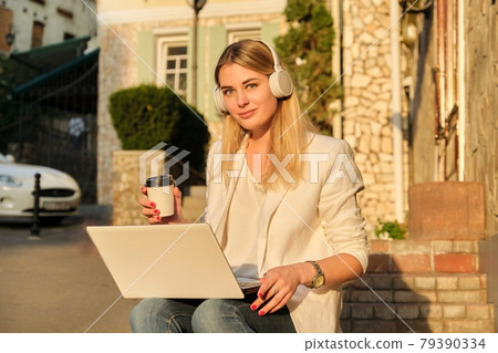 Young beautiful female in headphones with laptop drinking coffee in paper cup Young beautiful female in headphones with laptop drinking coffee in paper cup 79390334